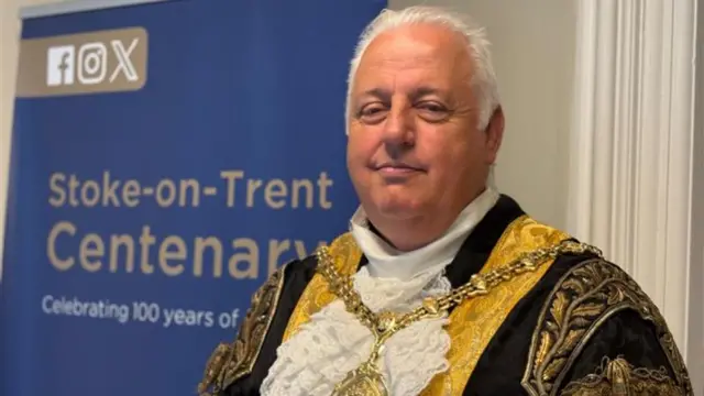 A man with short white hair, wearing gold and black mayoral robes with a gold chain around his shoulders. Behind him is a blue banner that says "Stoke-on-Trent Centenary".