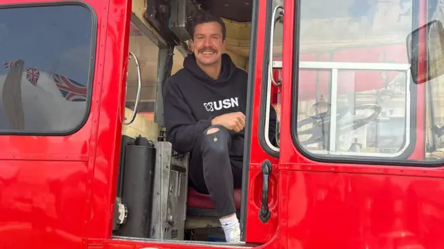 A man wearing a black hoody and black trousers, with white trainers, smiles while sitting aboard a red bus.