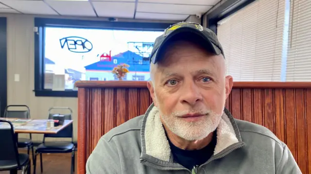 A man looks at a camera, in a restaurant, with a cap.
