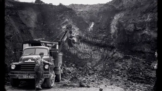 An old black and white image taken from the bottom of a large pit. A man in worker's overalls is leaning against the front of an old truck.
