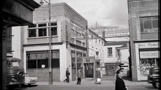 A black and white image showing a high street with people walking and cars driving past. Between two buildings is a gated entrance with a metal sign above it that says "Spode Works".