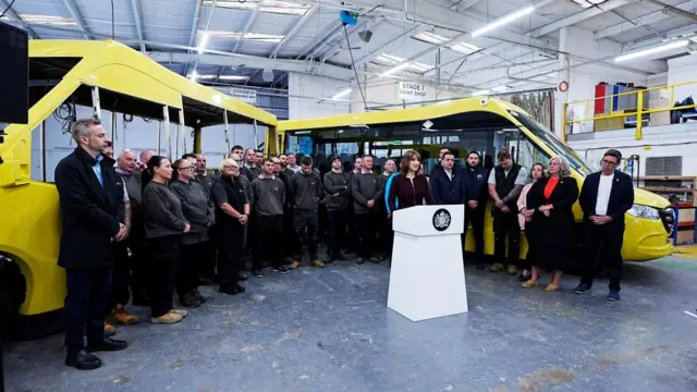Reeves delivers a speech in building, standing behind a white podium. Transport Sec Heidi Alexander and Mayor of Greater Manchester Andy Burnham stand to her left. Reeves is surrounding by workers and two buses.