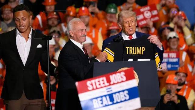 Trump is given a sport shirt at a rally which says his name on the back. A person hold a "steel jobs, real jobs" sign in the foreground