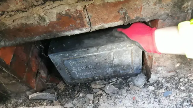 A black cuboid-shaped box in an opening in a wall. The box is old and damaged. It says "North Staffs Infirmary" on the front. Rubble is on the ground next to the box and a hand wearing a red glove has been placed on it.