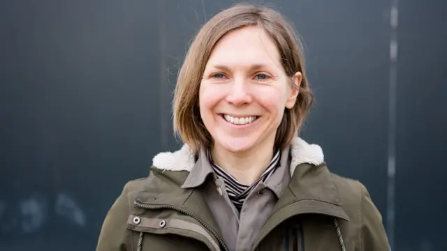 A woman with long brown hair and wearing a green coat over a grey jacket, smiles.