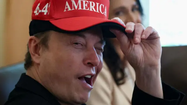 Musk winks at the camera as he tips his Maga cap