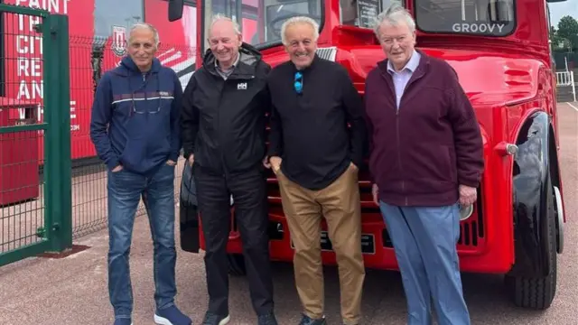 Four elderly men standing in front of a red bus parked next to a football stadium.