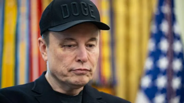 Musk, wearing a black cap that reads DOGE, looks down