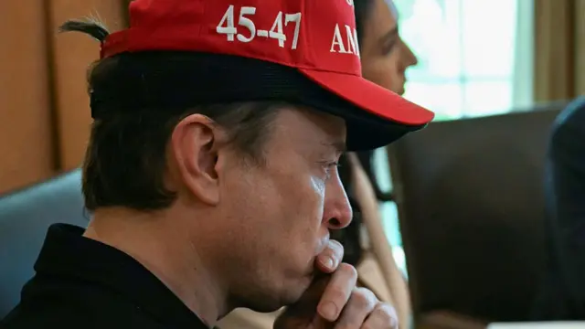Elon Musk wears two caps, and looks to he right of frame, his hand on his chin with a pensive look on his face