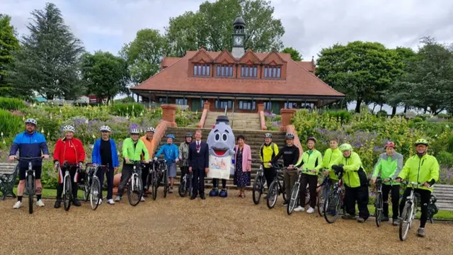 A group of people, most of whom are holding bicycles, line up and pose for a picture with a mascot of a grey bottle oven in the middle. They are standing on a stone-covered courtyard at the bottom of some steps leading up to a building.