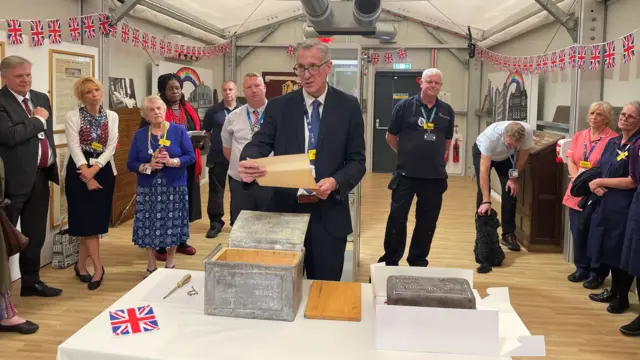 A man stands next to an old, silver box, holding a letter, which has been yellowed by time. He appears to be reading out what the letter says while other people watch on.