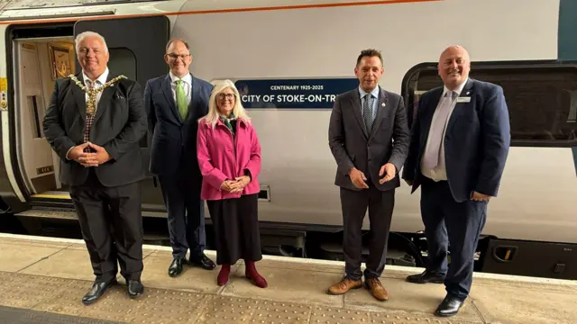 Four men and a woman stand on a platform next to a train with an open door. On the side of the train is a blue sign that says "Centenary 1925-2025, City of Stoke-on-Trent".