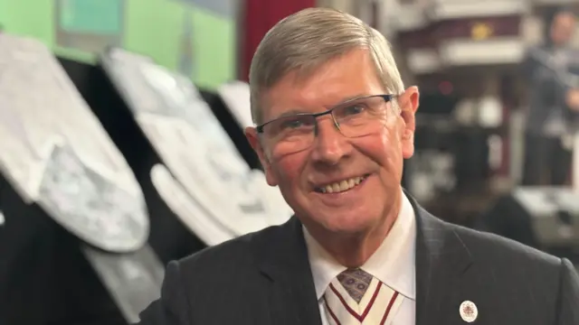 A man with short grey hair, glasses, a white shirt and black jacket, smiles at the camera.