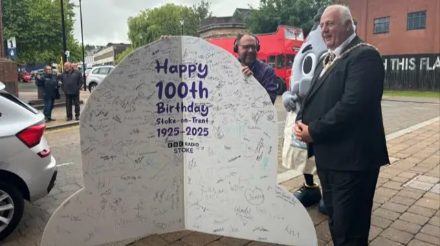 An massive open birthday card being presented to a man in a suit wearing a gold mayoral chain. The card is covered in signatures and says "Happy 100th Birthday Stoke-on-Trent 1925-2025".