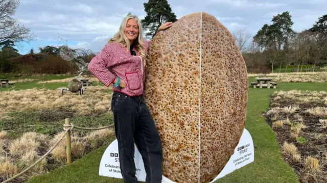 A woman wearing a red patterned jacket and black jeans is stood in an ornamental garden next to a card which is the same height as her. The card resembles a traditional Staffordshire oatcake.