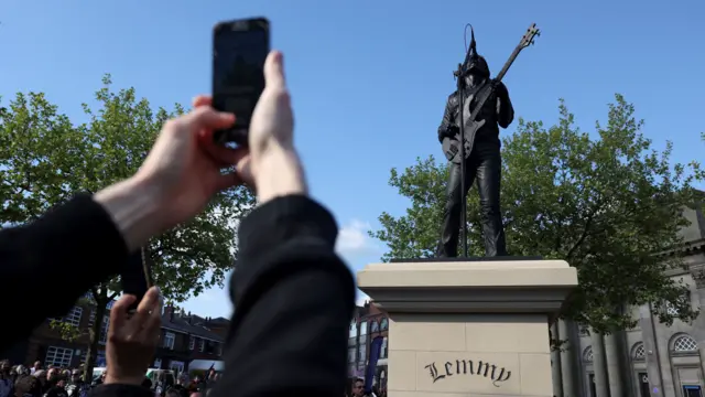 A person holds up a phone with both hands as they take a picture of the statue of Lemmy Kilmister in Burslem, which is placed on a plinth that says "Lemmy".