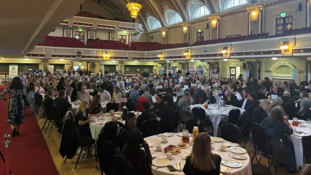 A large number of people sitting at round tables with white table clothes, in a big hall with hanging lights from the ceiling.