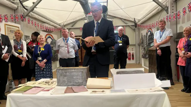 A man stands behind a table with an old silver box, surrounded by documents. The man is reading from a pamphlet while others watch on.