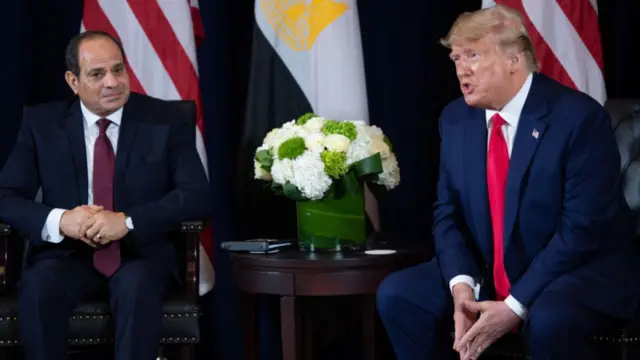 Trump and el-Sisi sit beside each other, separated by a small wooden table. They are both wearing navy suits and red ties, and Trump's mouth is open as he speaks.