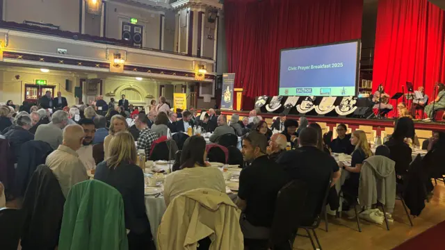 A large number of people sitting at round tables with white table clothes in a big hall. A stage is in the background with red curtains at the back and a large screen at the front which says "Civic Prayer Breakfast 2025".