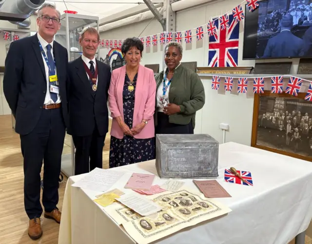 Two men and two women stand next to a table, on which is an old silver box with old documents laid out next to it. Behind them, the wall is covered in Union Flag bunting.