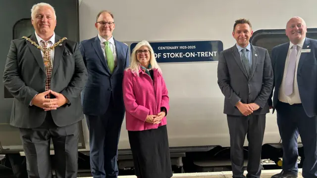 Four men and a woman stand on a platform next to a silver train which has a blue sign on it that says "Centenary 1925-2025 City of Stoke-on-Trent".