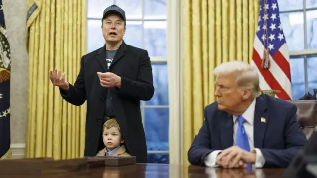 Donald Trump sits at the Oval Office desk. Musk, standing behind his son, makes gestures as he speaks to the media.