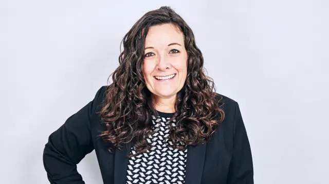 A woman, wearing a black blazer and black and white striped top, smiles with a hand on her hip.