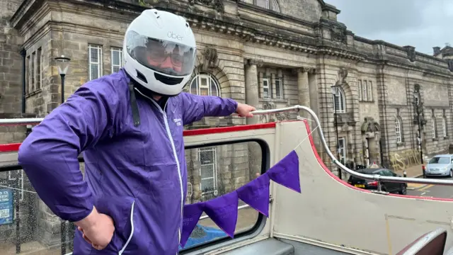 A man wearing a white crash helmet and a purple jacket stands on top of an open-topped bus with his right hand on his hip and his left hand resting on a rail.