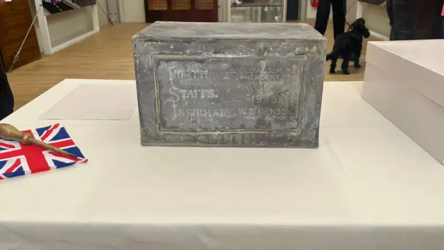 An old grey box which says North Staffs Infirmary, placed on a table covered with a white cloth. A small union flag and screwdriver have been placed to the side of it.