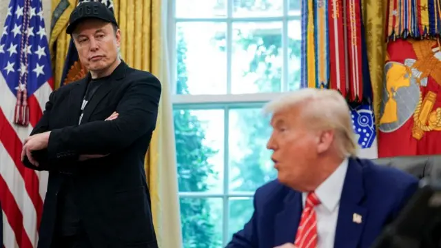 Musk with his arms crossed in the Oval Office, looking over to Trump sat on the Oval Office desk