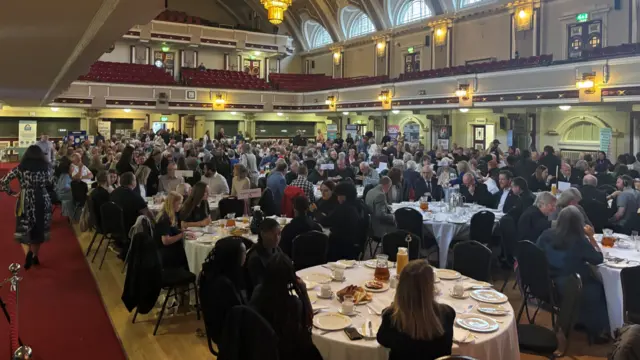 A large number of people sitting around circular white tables in a hall.