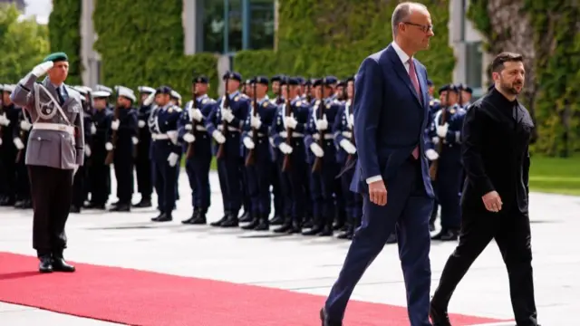 Zelensky and Merz walking together on a red carpet in front of a military parade
