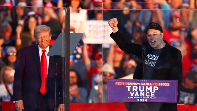 At at Trump re-election rally, Musk raises his fist at a podium standing next to Donald Trump.