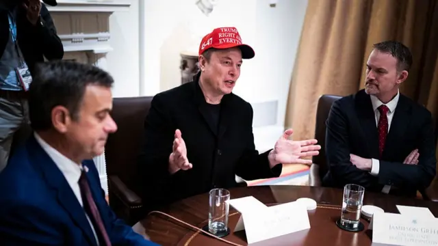Elon Musk speaking, hands raised whether a Trump hat. Two men sat beside him.