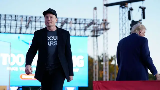 Musk walking towards camera, Trump stood behind him on stage, with back to camera.