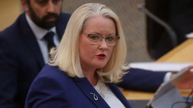Christina McKelvie, a woman with bright blonde hair. She is wearing glasses, red lipstick and a blue blazer jacket.