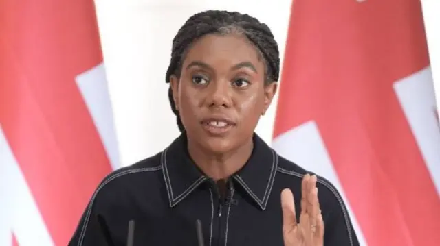 Kemi Badenoch, wearing a dark denim shirt, her braided hair is tied back. She is standing at a podium in front of several union jacks.