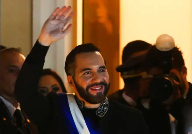 Nayib Bukele waving to someone in the distance with a press photographer taking a photo behind him