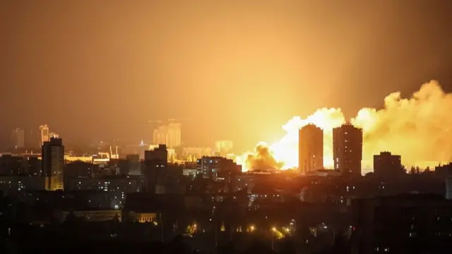 Wide shot of Kyiv skyline in the dark, illuminated by a bright orange flash behind some buildings, with a plume of white smoke.