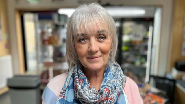 Norma Hastie looks smiling into the camera, wearing a blue scarf and a pink top