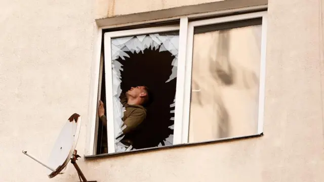 A man is seen, through a smashed apartment window above a satellite dish, inspecting the inside of a building