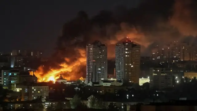 Wide shot of buildings in Kyiv in the dark, with a bright orange flame and grey smoke billowing out among the buildings