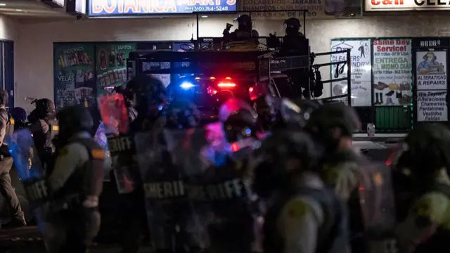Law enforcement officers wearing helmets and carrying shields walk past shopfronts, while two officers are seen sat on a truck that has its police lights on