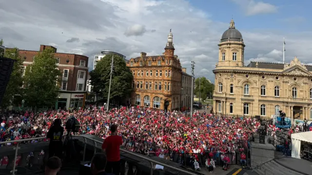 Queen Victoria Square in Hull looking towards the maritime museum and other buildings. A large crowd is gathered around a central statue in the square