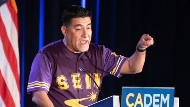 David Huerta speaking on stage wearing a purple and yellow sport jersey, a lectern in front of him carrying a sign reading CADEM
