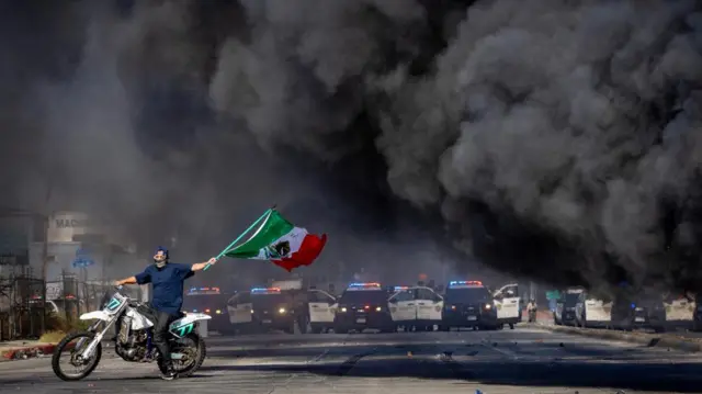 A man, wearing a covering on the lower part of his face, is seen waving a Mexican flag while on a motorbike as police cars are seen lined up in the distance