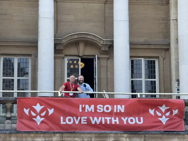 Pinkney stands on the left of the picture with James Hoggarth on the balcony above a banner that reads "I'm so in love with you"