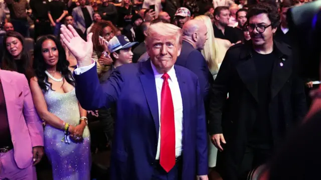 Donald Trump in stadium, hand in the air and smiling, large crowds behind him. He's in a blue suit with long red tie.