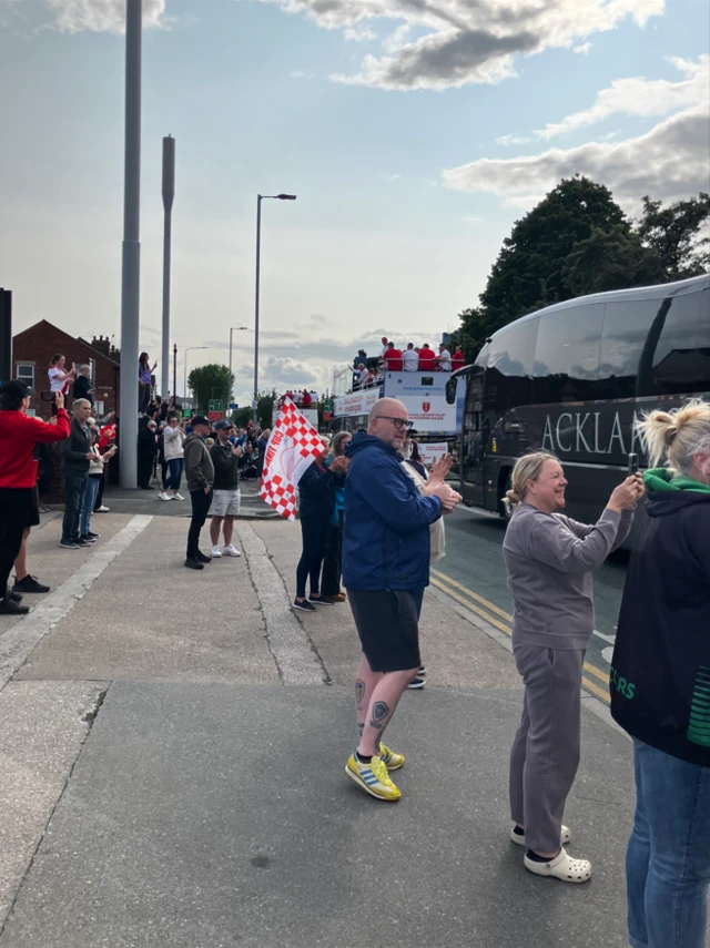 People clap and cheer as an open-top bus passes them closely followed by a grey and silver coach